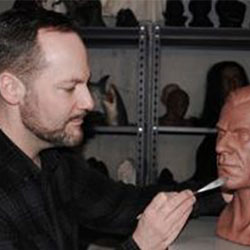A man sculpting details on a human head bust with a tool in front of shelves displaying other busts.