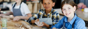 Happy little girl looking at you while sitting by table and making clay cup with her schoolmates on background