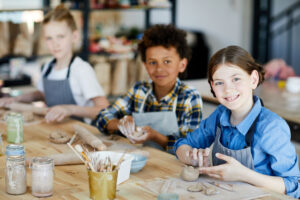 Happy little girl looking at you while sitting by table and making clay cup with her schoolmates on background