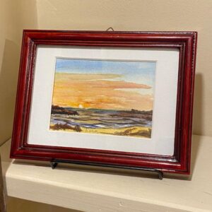 Afterglow" by Diane Orzel, a framed sunset landscape watercolor, displayed on a small stand against a beige wall.