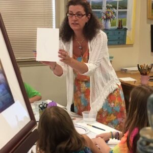 A woman holds a blank sheet as kids with art supplies sit at a table in the Friday Homeschool Teen Art Class with Kristine Mancuso.