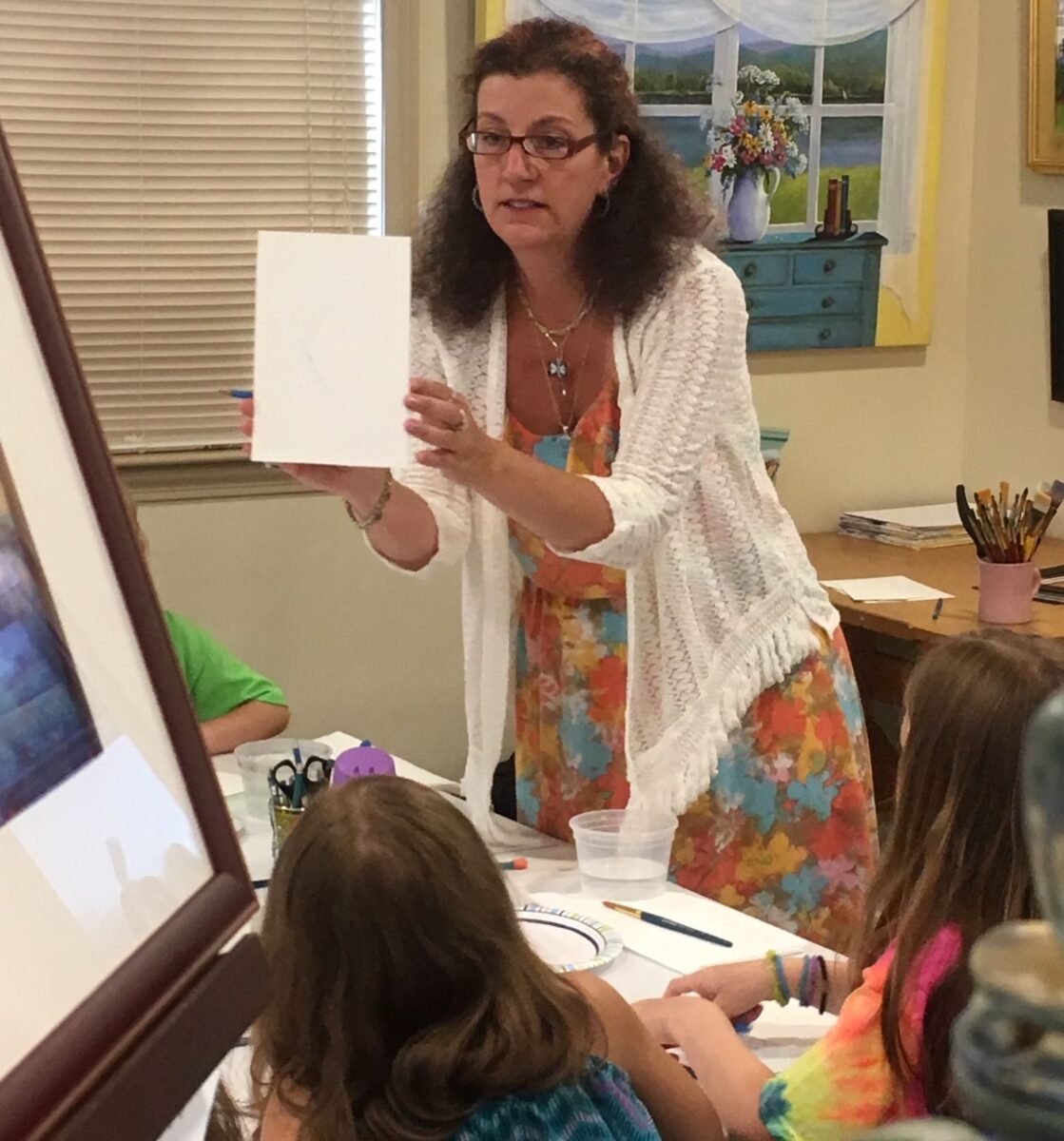 A woman holds a drawing as kids with art supplies sit at a table in the Friday Homeschool Teen Art Class with Kristine Mancuso.
