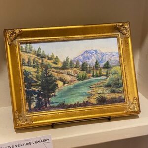 Mt. Views" by Toni DeRuntz: a framed painting of a river and mountains, displayed on a shelf.