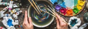 colorful showing artist hands swirling colorful paint in a bowl surrounded by supplies and tubes...